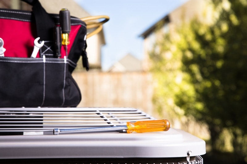 Blog Title: Common AC Problems and Solutions Photo: Bag of repairman's work tools, screwdriver on top of air conditioner unit outside brick home.